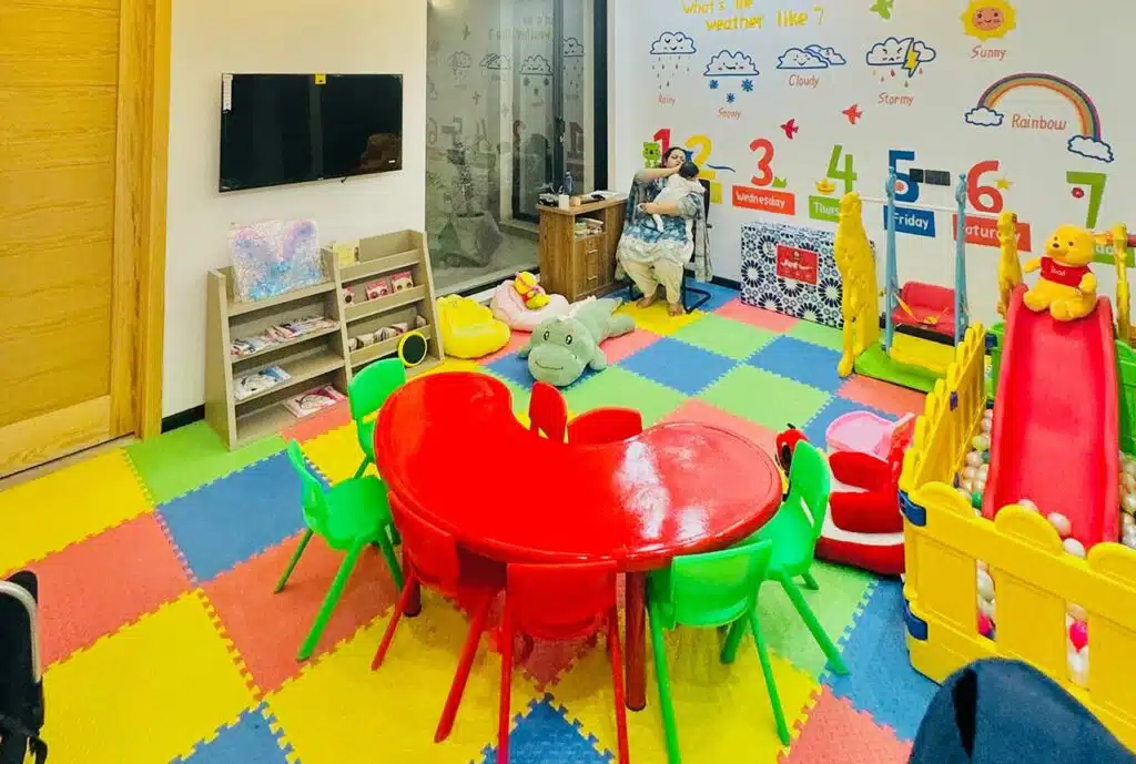 Brightly colored children's playroom with a red table and green chairs, featuring toys, a slide, and a play area with foam flooring. A child-sized bookshelf and a television are visible in the background.
